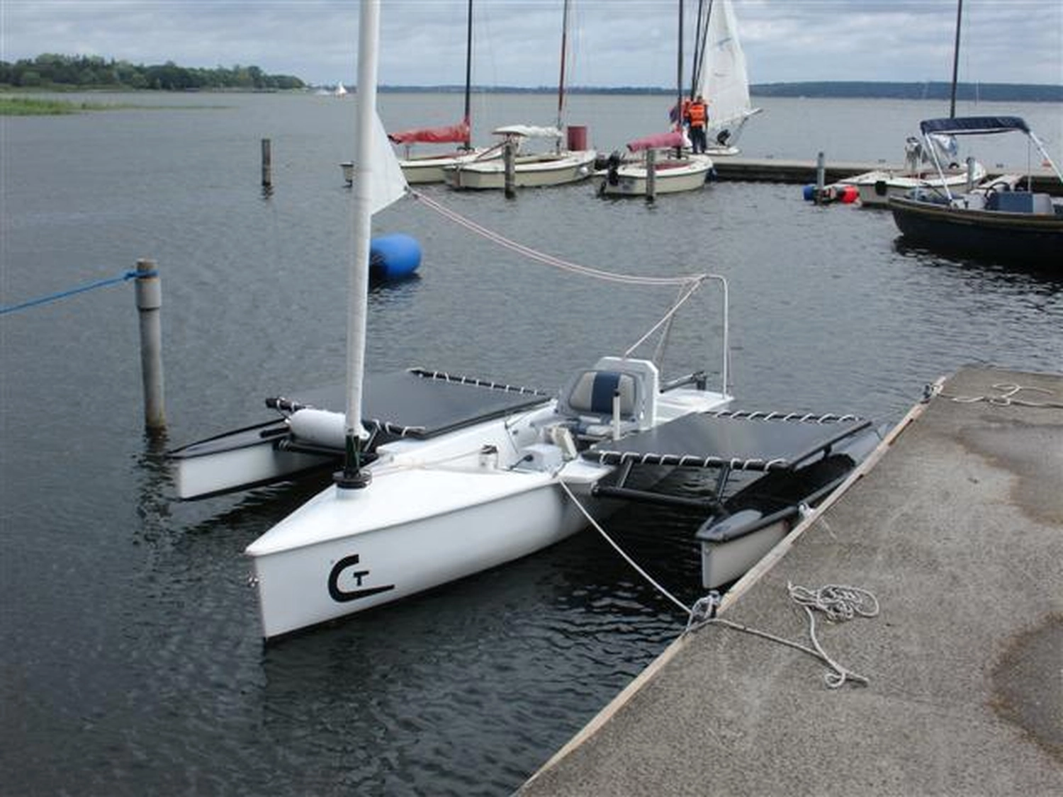 HCS Handels Contor Schönwälder GmbH - Innovative Techniklösungen für Energie, Wasser und Mobilität - Ein kleines, weißes Trimaran-Segelboot mit Muskelkraftantrieb liegt an einem Pier vor Anker.