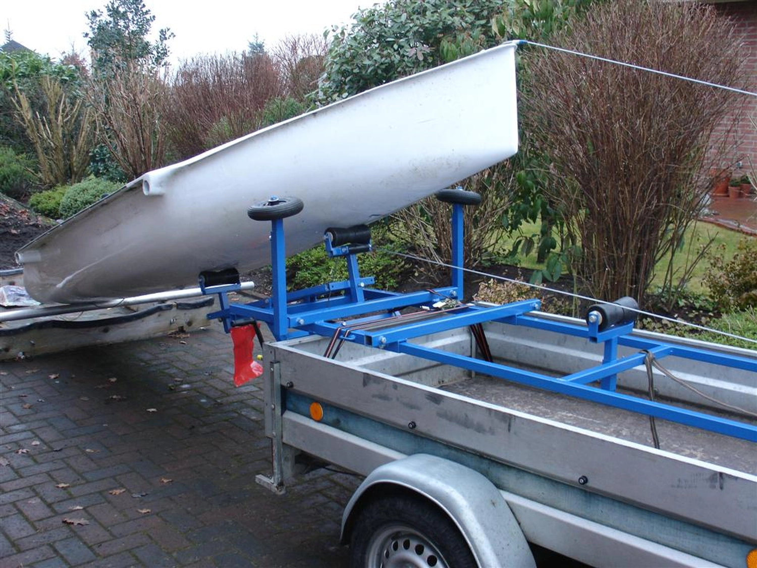 HCS Handels Contor Schönwälder GmbH - Innovative Techniklösungen für Energie, Wasser und Mobilität - Ein weißes Flying Fifteen-Segelboot steht auf einem blauen Slipwagen, der an einem silbernen Straßenanhänger befestigt ist. Das Bild zeigt die Vorbereitung des Bootes zum Zuwasserlassen oder Bergen; das Heck des Bootes ruht auf den Rädern des Slipwagens. Im Hintergrund ist eine bewachsene Freifläche zu sehen.