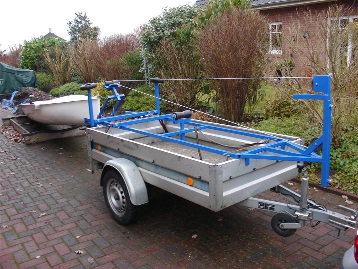 HCS Handels Contor Schönwälder GmbH - Innovative Techniklösungen für Energie, Wasser und Mobilität - Ein weißes Flying Fifteen-Segelboot steht auf einem blauen Slipwagen, der an einem silbernen Straßenanhänger befestigt ist. Das Bild zeigt die Vorbereitung des Bootes zum Zuwasserlassen oder Bergen; das Heck des Bootes ruht auf den Rädern des Slipwagens. Im Hintergrund ist eine bewachsene Freifläche zu sehen.