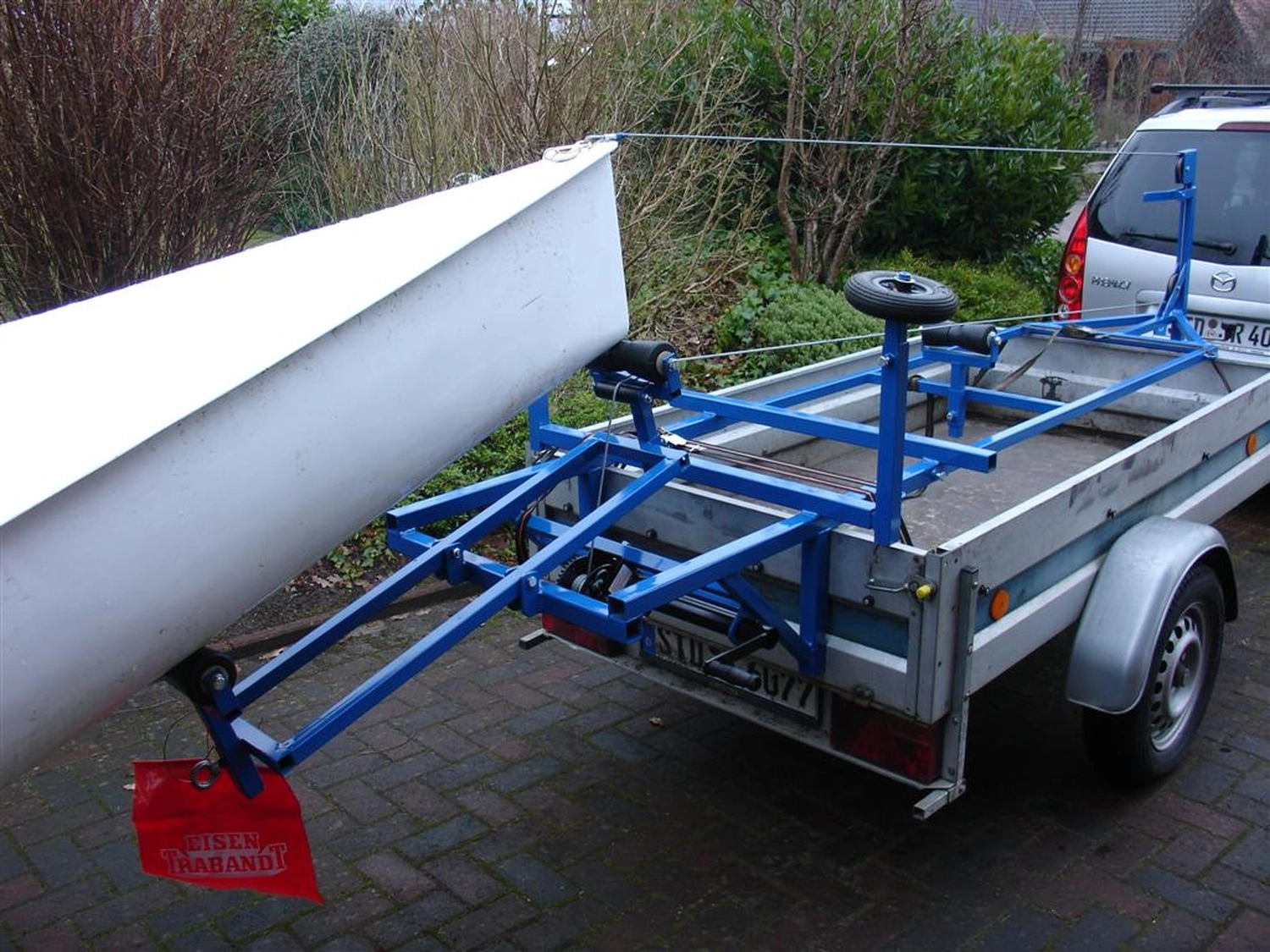 HCS Handels Contor Schönwälder GmbH - Innovative Techniklösungen für Energie, Wasser und Mobilität - Ein weißes Flying Fifteen-Segelboot steht auf einem blauen Slipwagen, der an einem silbernen Straßenanhänger befestigt ist. Das Bild zeigt die Vorbereitung des Bootes zum Zuwasserlassen oder Bergen; das Heck des Bootes ruht auf den Rädern des Slipwagens. Im Hintergrund ist eine bewachsene Freifläche zu sehen.