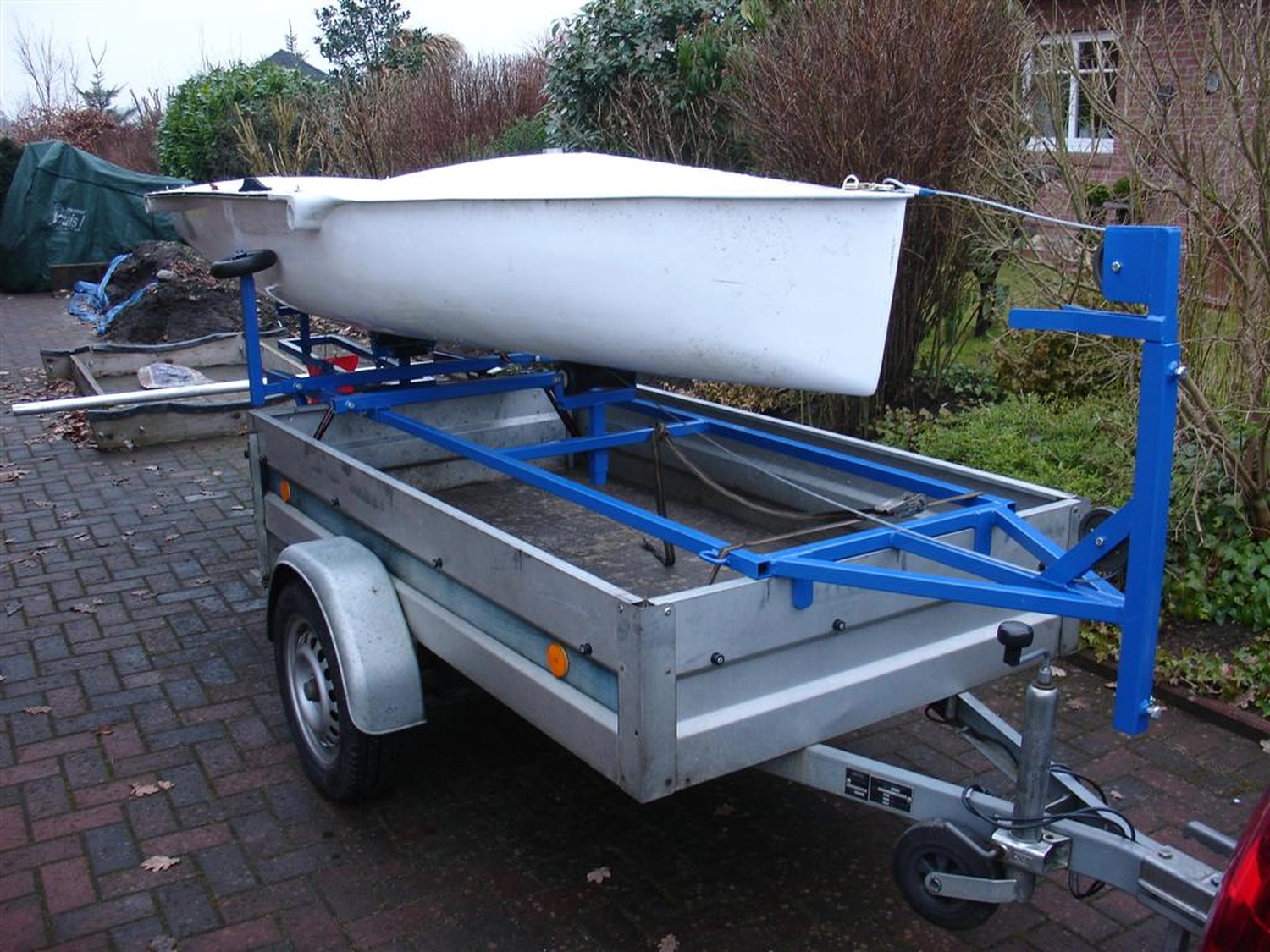 HCS Handels Contor Schönwälder GmbH - Innovative Techniklösungen für Energie, Wasser und Mobilität - Ein weißes Flying Fifteen-Segelboot steht auf einem blauen Slipwagen, der an einem silbernen Straßenanhänger befestigt ist. Das Bild zeigt die Vorbereitung des Bootes zum Zuwasserlassen oder Bergen; das Heck des Bootes ruht auf den Rädern des Slipwagens. Im Hintergrund ist eine bewachsene Freifläche zu sehen.