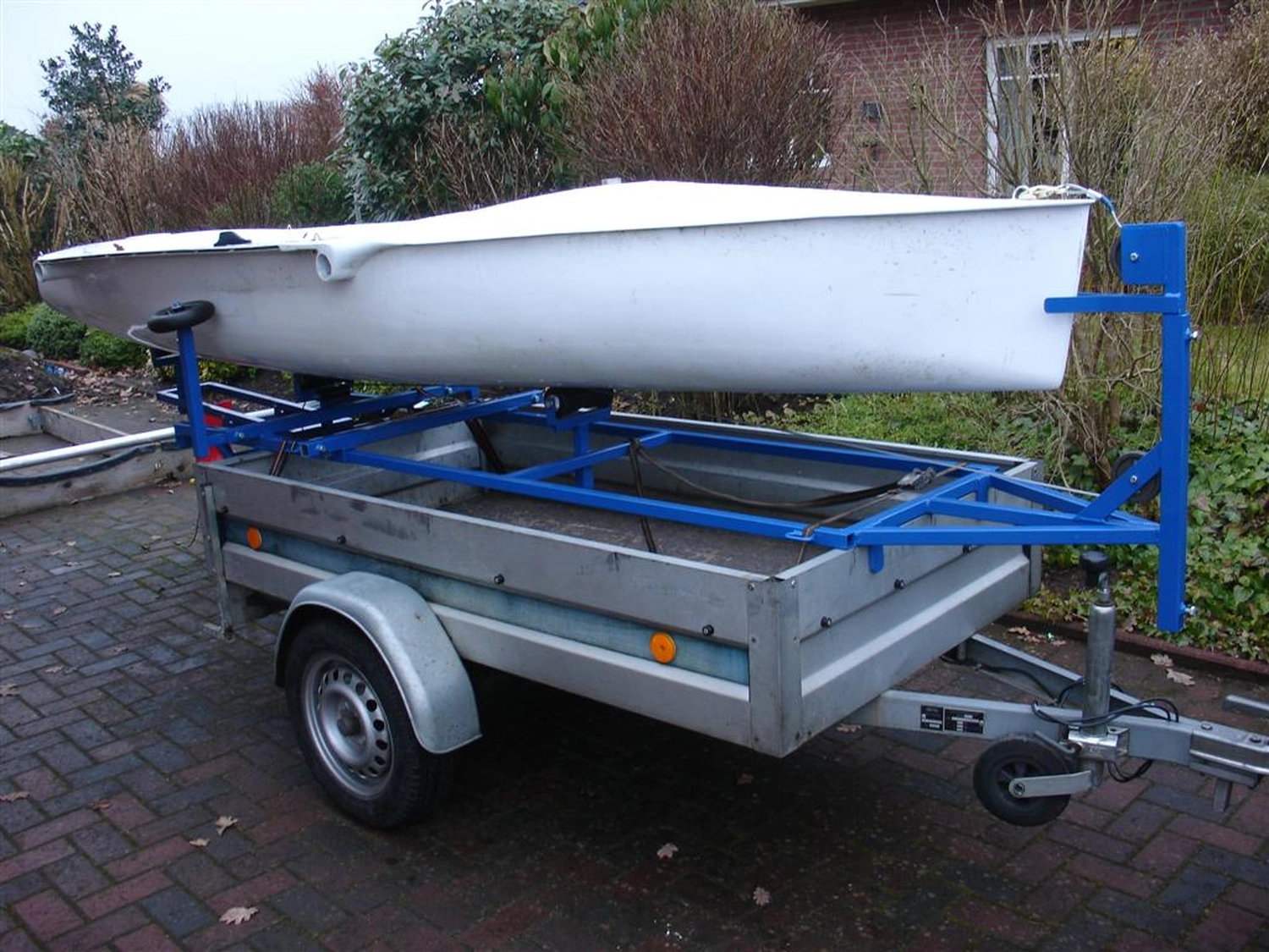 HCS Handels Contor Schönwälder GmbH - Innovative Techniklösungen für Energie, Wasser und Mobilität - Ein weißes Flying Fifteen-Segelboot steht auf einem blauen Slipwagen, der an einem silbernen Straßenanhänger befestigt ist. Das Bild zeigt die Vorbereitung des Bootes zum Zuwasserlassen oder Bergen; das Heck des Bootes ruht auf den Rädern des Slipwagens. Im Hintergrund ist eine bewachsene Freifläche zu sehen.