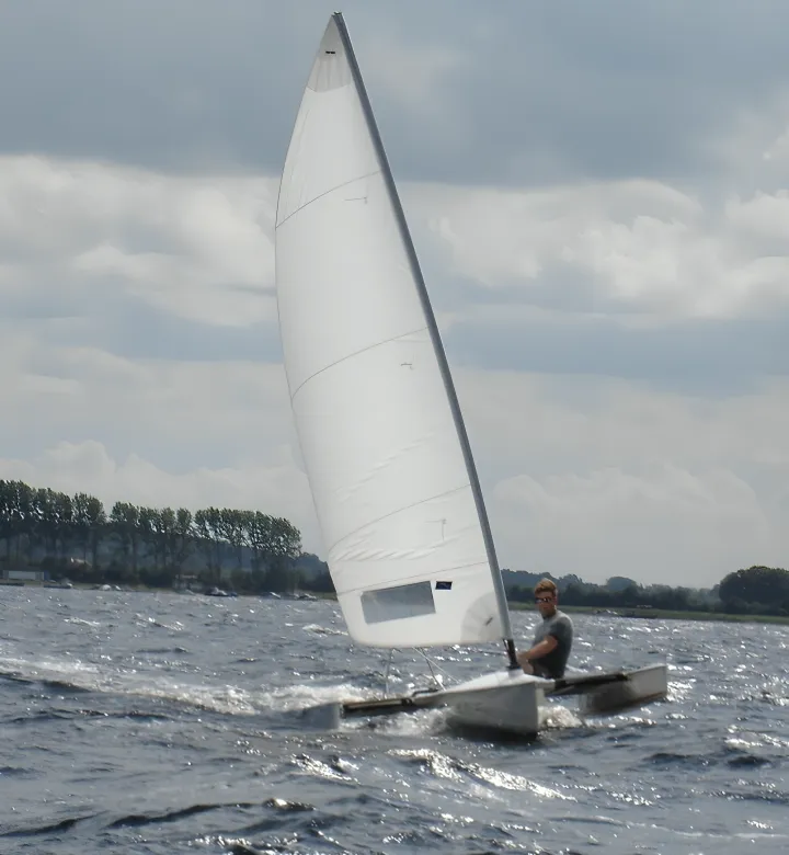 HCS Handels Contor Schönwälder GmbH - Innovative Techniklösungen für Energie, Wasser und Mobilität - Eine Person segelt in einem kleinen, weißen Viola 14 Segelkanu auf unruhigem Wasser unter bewölktem Himmel. Das Boot ist ein leichtes, leistungsstarkes Segelkanu mit einem stabilen Gleitrumpf und einem einzelnen weißen Segel. Der Segler sitzt im Rumpf, und am fernen Ufer sind Bäume zu sehen.